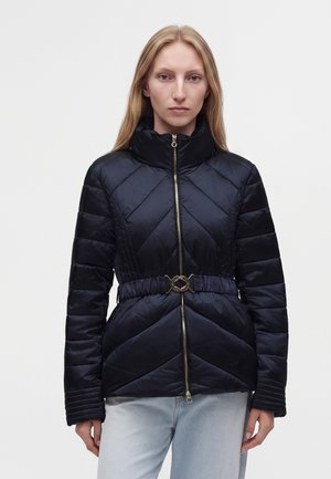 Femme aux cheveux blonds et raides portant une veste matelassée noire avec un col montant, une fermeture éclair devant et une ceinture à la taille, associée à un jean bleu clair.
