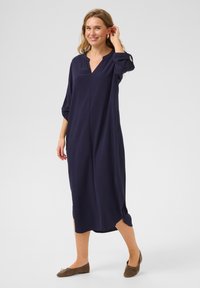 Mörkblå, knälång klänning med V-ringning, tre fjärdedelars ärmar, sidoflikar och lätt tyg. Bärs med bruna ballerinas.