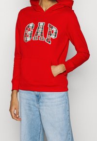 Sudadera roja con capucha y bolsillo frontal, con letras de cuadros que forman la palabra "GAP". Mangas largas y capucha con cordón completan el diseño casual.