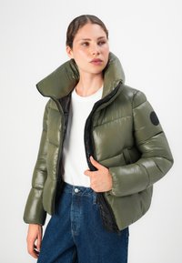 Vrouw in een groene pufferjack met hoge kraag, witte shirt en blauwe jeans, die de rits van de jack vasthoudt terwijl ze opzij kijkt.