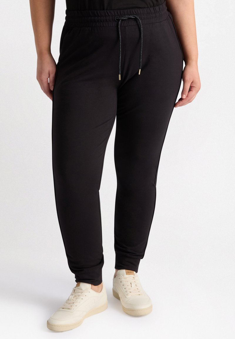 Schwarze, schmal zulaufende Jogginghose aus weichem Stoff, mit einem elastischen Bund und einer Kordel zum Verstellen, kombiniert mit hellen Sneakern.