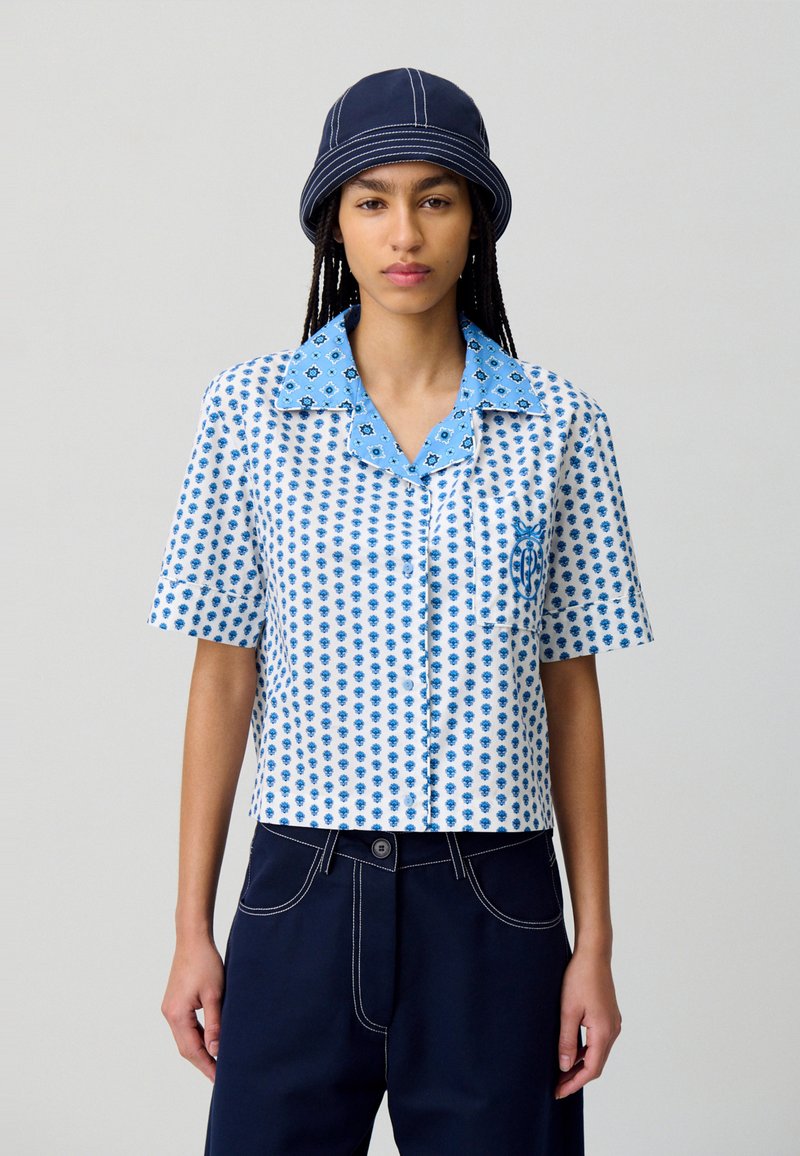 Camicia a maniche corte bianca con motivo floreale blu e colletto blu a contrasto. Accostata a pantaloni blu navy con dettagli di cucitura bianchi.