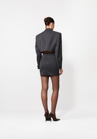 Kobieta z włosami związanymi w kok, ubrana w ciemnoszary, krótką marynarkę, dopasowaną mini spódnicę, przejrzyste czarne rajstopy i czarne szpilki, stojąca na białym tle.