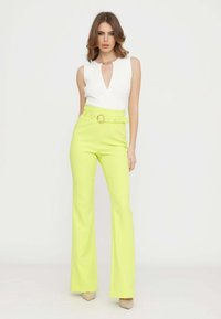 Body bianco a coste con scollatura a V profonda e dettaglio a catena, abbinato a pantaloni a palazzo giallo neon a vita alta e tacchi beige.