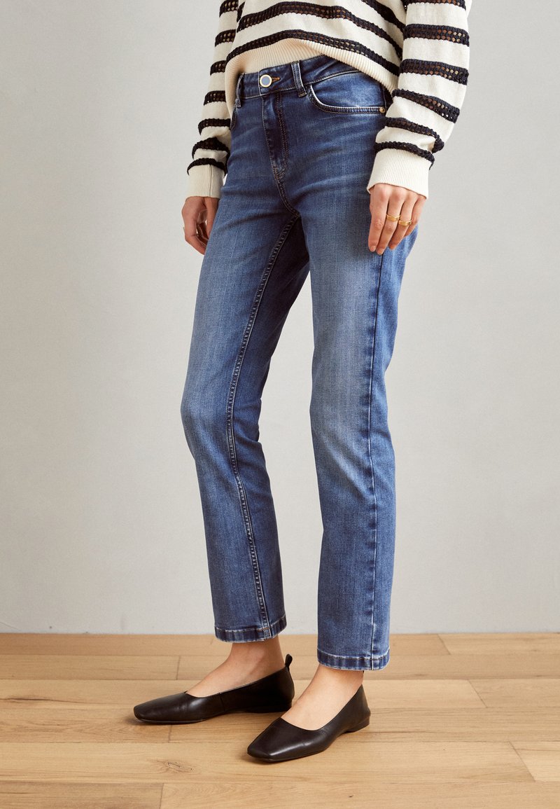 Mørkeblå, tettsittende jeans med middels høy midje, falmede detaljer og cropped lengde, kombinert med svarte flate sko på lys, tregulv.
