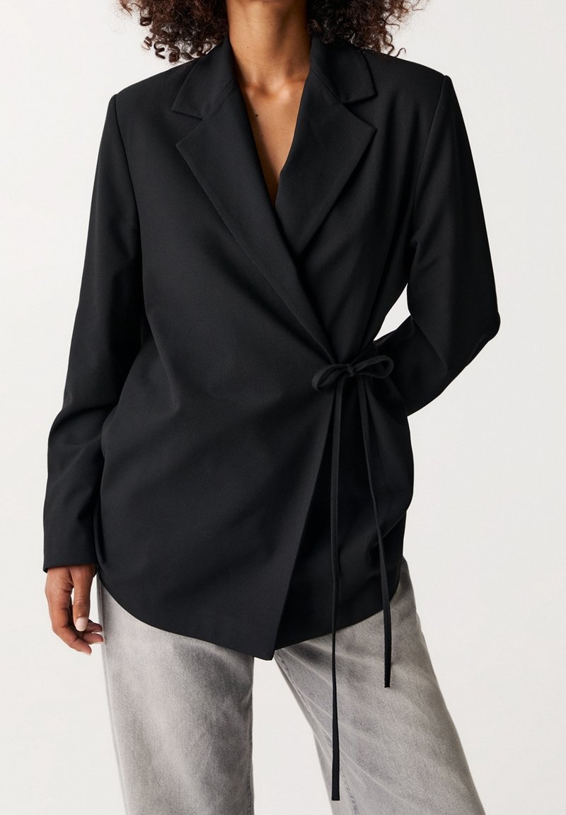 Blazer noir portefeuille en tissu lisse, avec ceinture à la taille, revers aiguisés et manches longues, associé à un pantalon gris clair.