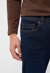 Prawa ręka obok kieszeni ciemnoniebieskich dżinsów, osoba w brązowej koszuli z długim rękawem, zbliżenie na dolną część ciała.