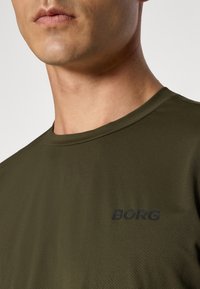 Oliven grønn kortermet skjorte laget av teksturert materiale, med en svart logo "BORG" på brystet. Enkel design og rund hals.