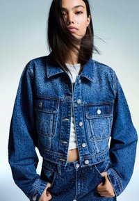 Denim skraćena jakna s teksturiranom površinom i ukrasima od perlica, dva prednja džepa i srebrnim detaljima. Boja: tamnoplava.