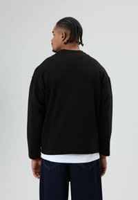 Zwarte sweat met lange mouwen en een ontspannen pasvorm. Gemaakt van een zachte stof, met verzakte schouders en een gelaagd wit overhemd eronder.