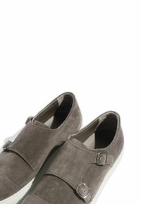 Sive suede natikače s dva podesiva metalna kopčanja, zaobljenim vrhom i bijelim gumeni potplat. Glatka tekstura i minimalistički dizajn.