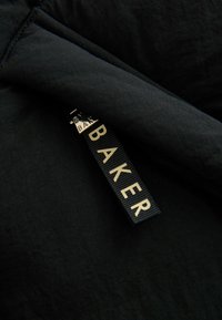 Tessuto nero che mostra un primer piano di un'etichetta con la scritta "BAKER" in lettering dorato su uno sfondo texturizzato. L'etichetta è attaccata al tessuto.