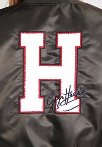 Svart satin bomberjacka med ett stort broderat vitt "H" som är konturerat i rött, med en signatur i blått i botten.