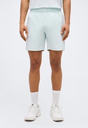 Uomo che indossa pantaloncini sportivi azzurri chiari, calzini bianchi e scarpe da ginnastica bianche, in piedi contro uno sfondo bianco uniforme.