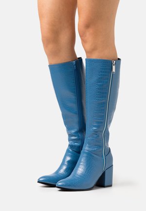 Bottes montantes en cuir synthétique bleu effet crocodile, avec un bout pointu, un talon bloc et une fermeture éclair latérale. Texture lisse avec une finition brillante.