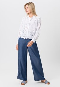 Witte oogjesblouse met bloemmotieven, lange mouwen en een golvende zoom, gedragen met losse donkere denim broek en blauwe slidesandalen.