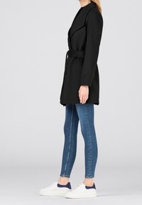 Cappotto nero con vita cintata, maniche lunghe e colletto a rever, abbinato a jeans skinny blu e scarpe da ginnastica bianche con dettagli blu navy.