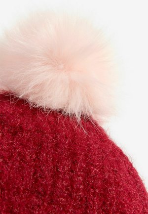 Červená pletená čiapka s mäkkým, nadýchaným svetloružovým brmbolcom na vrchu. Textúra je hustá a mierne chĺpkatá.