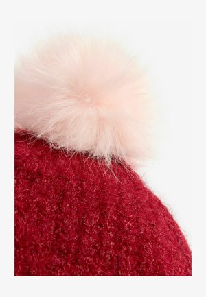 Sarkans adīts beanie ar mīkstu, pūkainu gaiši rozā pom-pom virsū. Tekstūra ir blīva un nedaudz pūkaina.