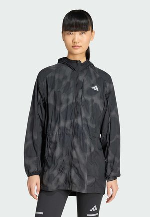 Kvinde med glat sort hår iført en sort mønstret hættetrøje og sorte leggings med hvide logoer, stående mod en ensfarvet baggrund.