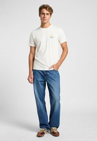 Camiseta de algodón blanca con un diseño gráfico colorido en el pecho izquierdo. Combinada con jeans de color azul claro y zapatillas marrones con acentos amarillos.