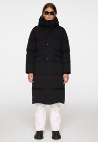 Svart puffjacka med hög krage, tryckknappar och sidofickor; har en quiltad struktur och är knälång. Bärs över vita byxor.