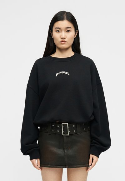 Νέα γυναίκα με μακριά σκούρα μαλλιά που φοράει μαύρο φούτερ "Palm Angels", μαύρη ζώνη με ασημένια αγκράφα και σκούρα μίνι φούστα σε απλό φόντο.