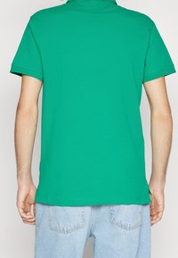 Polo vert avec une finition texturée, manches courtes et col classique ; associé à un jean bleu clair, montrant le détail de l'ourlet.