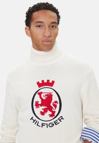 Suéter crema de cuello alto con escudo de león rojo y texto "HILFIGER", con cuello de canalé y puños a rayas azules. Textura de punto suave.