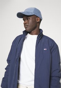 Marineblå zip-jakke med høj krave, båret over en hvid t-shirt. Blå corduroy-kasket, glat tekstur, og minimal branding-detalje synlig.
