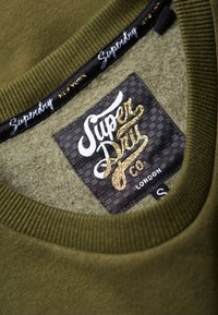 Cuello de sudadera verde oliva con borde acanalado, que presenta una etiqueta negra con texto dorado estilizado "Superdry Co" y un patrón de tablero de ajedrez.