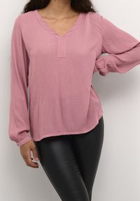 Blouse rose texturée avec col en V et détail plissé sur la poitrine. Manches longues avec poignets élastiques. Associée à un legging noir.