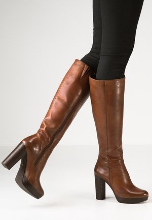 Bottes en cuir marron montantes jusqu'aux genoux avec des talons carrés hauts portées avec un pantalon noir sur un fond clair uni.
