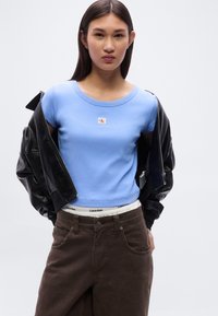 Ανοιχτό μπλε κοντομάνικο t-shirt με στρογγυλή λαιμόκοψη, μαύρο δερμάτινο μπουφάν bomber και καφέ παντελόνι ψηλόμεσο με ζώνη Calvin Klein.