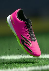 Lys rosa fotballsko med svarte lisser og logo, limegrønn såle, i luften over en gressbane med små gresspartikler som flyr rundt.