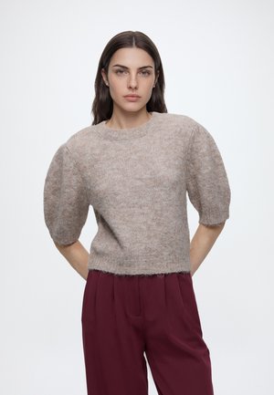 Kvinde med glat mørkt hår iført beige, blød kortærmet sweater og højttaljede vinrøde bukser, stående med hænderne bag ryggen.