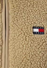 Béžová fleecová bunda s texturovaným povrchem, vybavená zipovým zapínáním a malým vyšitým logem "Tommy Jeans" v modré, červené a bílé barvě.