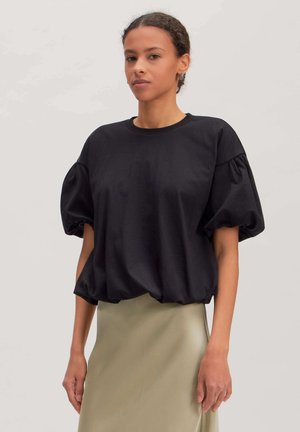 Woman with short curly hair wearing a black puff sleeve top and beige skirt standing against a plain light background.