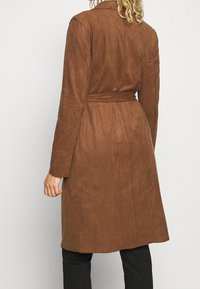 Manteau en daim marron avec une taille ceinturée, des manches longues et un col simple ; texture lisse et design mi-long avec une coupe droite.