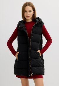 Chaleco de plumas negro con capucha, con herrajes dorados, textura acolchada horizontal y bolsillos laterales, usado sobre una camiseta de manga larga roja.