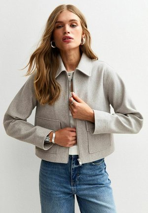 Ljusgrå cropped jacka i mjukt material med krage, dragkedjestängning och två framfickor; bärs över en vit topp och blå jeans.