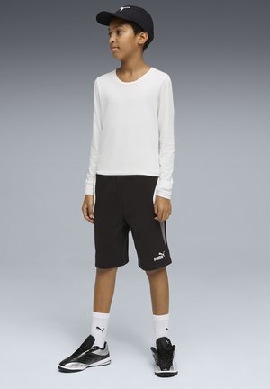 Jongen staand, draagt een zwarte Puma-pet, wit shirt met lange mouwen, zwarte Puma-short, witte Puma-sokken en zwarte en zilveren Puma-sneakers.