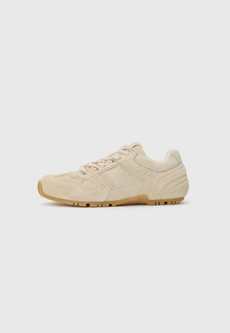 Beige suede sneaker with textured accents, rounded toe, light-colored laces, and a gum rubber sole. Features perforated detailing on the sides.