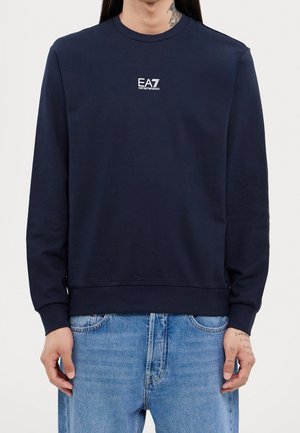 Person trägt einen marineblauen EA7 Emporio Armani Sweatshirt mit langen Ärmeln und hellblaue Jeans, zeigt Hand- und Hals-Tattoos.