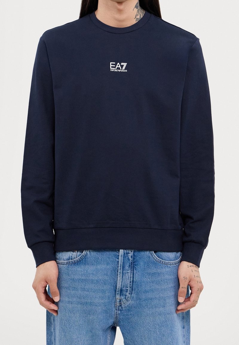 Personne portant un sweat-shirt à manches longues bleu marine EA7 Emporio Armani et un jean bleu clair, montrant des tatouages aux mains et un tatouage au cou.