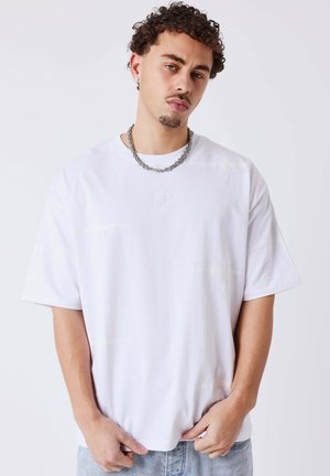 Jonge man met krullend haar draagt een eenvoudige witte oversized T-shirt, een ketting en lichtblauwe spijkerbroek, staand tegen een witte achtergrond.