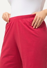 Rode katoenen broek met een elastische tailleband, voorzien van zijzakken en een gladde textuur, gedeeltelijk zichtbaar op taillehoogte.