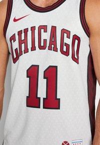 Biała koszulka koszykarska z czerwonym napisem "CHICAGO" i numerem "11". Teksturowany wzór diamentowy z czarnymi i czerwonym wykończeniami. Wyświetlone logo Nike.