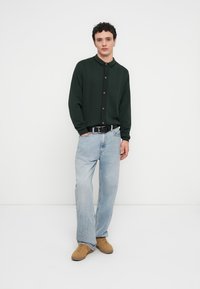 Chemise en tricot vert foncé boutonnée avec col, associée à un jean large bleu clair et des chaussures marron clair. Design minimaliste sur fond blanc.