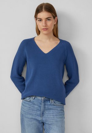 Blauwe V-halstrui met een geribbelde textuur en lange mouwen, gecombineerd met lichte denim jeans. Het materiaal lijkt zacht en aansluitend.